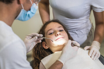 High angle shot of a young girl laying calm in the dental chair, looking at the camera and smiling. The dentist and his assistant prepared to examine her teeth. - Powered by Adobe