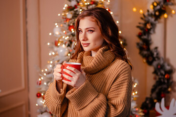 Close-up portrait of an attractive young woman wearing a warm sweater with a red mug in her hands on the background of a Christmas tree. The girl drinks a drink in a cozy New Year's interior.