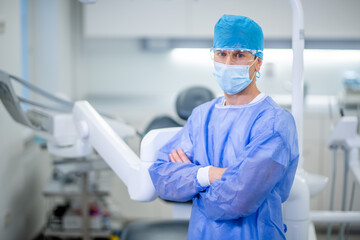 Portrait of a Caucasian male dentist standing with crossed arms in a dental clinic. The dentist with a protective face mask, safety glasses, and a blue surgical cap.