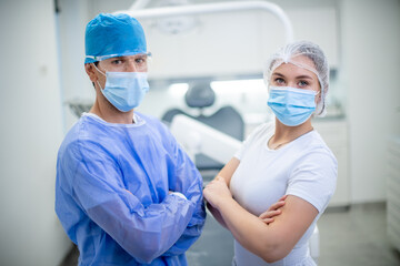 Portrait of a Caucasian male dentist and his female assistant in a dental clinic. The dentist wearing safety glasses, a protective face mask, and a blue surgical cap.
