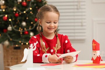Cute little girl cutting paper at table with Saint Nicholas toy indoors