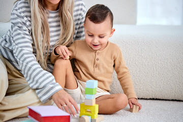 Obraz premium Smiling caucasian lovely mother play with little kid son sit on warm floor, caring mom nanny help teach child boy build constructor of colorful blocks at home, daycare, children development