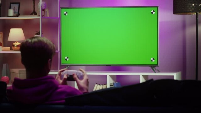 Man playing video game in living room in evening. Young guy sitting on sofa against tv with chroma green screen. Student gaming with modern console.