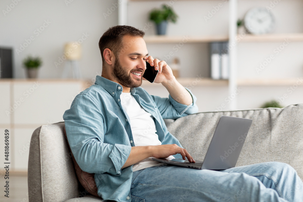 Poster attractive young guy talking on smartphone and using laptop computer on couch at home office