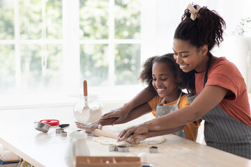 Cheerful black mother helping daughter rolling pasrty dough