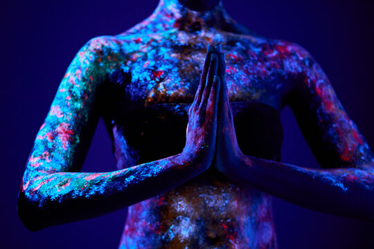 cropped slender sensual woman of black appearance in fluorescent paint makeup, posing in prayer pose. luminescence paint, body art, neon lights. isolated on dark background