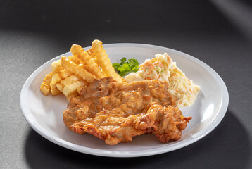 Deep fried breast chicken cutlet with french fries and salad in white dish on dark grey background top view
