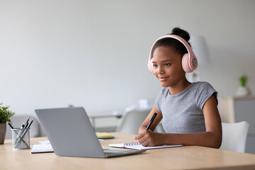 Happy adolescent afro american female student with headphones studying at home watching online lesson