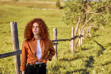 Portrait Of Adorable Redhead Lady In Casual Orange Shirt And Black Trousers Posing At Camera In Field In Countryside, Looking Calm And Relaxed, in Contemplation. Curly Freckled Woman At Sunset