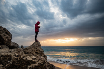 Fototapeta na wymiar Young man standing on a rock watching waves breaking at the Cape of Trafalgar in a dramatic stormy sunset 