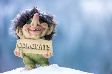 Cute happy troll holding sign with the word congrats chiseled out. Soft blurred out background in...