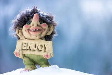 Cute happy troll holding sign with the word enjoy chiseled out. Soft blurred out background in winter wonderland.