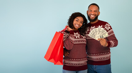Happy african american spouses in Christmas sweaters holding shopping bags and bunch of money over blue background