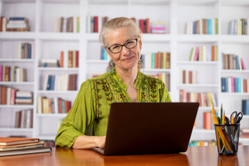 Fototapeta premium Portrait of smiling woman with glasses looking at camera in home office with library or books background.