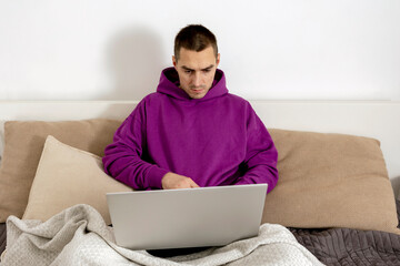Young caucasian man with violet hoodie sitting on bed and holding laptop computer. Man using notebook to surf in internet, read news, watch movie, study or work online. Relaxing at home.