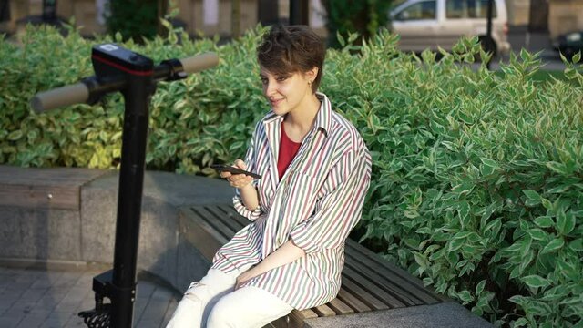 Charming relaxed young woman sitting in sunbeam on city street surfing Internet on smartphone smiling looking around. Portrait of happy carefree Caucasian lady enjoying leisure outdoors in sunshine