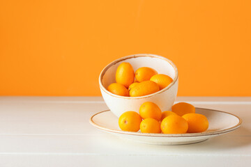 Bowl and plate with tasty kumquat fruits on table against orange wall