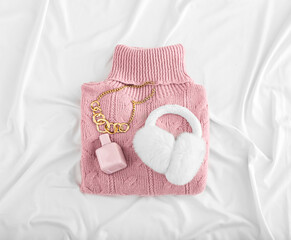 Knitted roll-neck sweater, perfume bottle and ear muffs on light crumpled fabric background