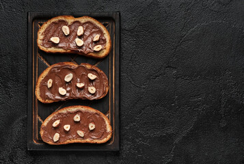 Wooden board of bread with chocolate paste and hazelnuts on black background