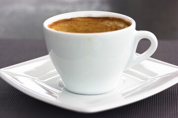 White cup of coffee. Round ceramic cup with cappuccino latte with brown foam on square saucer on a table. A hot flavored aromatic drink in a cafe, restaurant in the morning, lunch. Beverage theme.