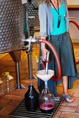 As if pumping gas, a woman fills a glass jug with wine from a nozzle and hose attached to a vat, in...