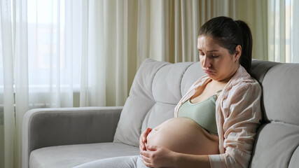 Pregnant upset brunette woman in flannel shirt and top sits on sofa of living room and strokes...