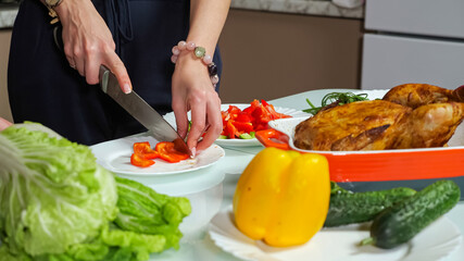 Housewife hands cook dinner with baked chicken cutting fresh red bell pepper for salad on plate near vegetables on table in kitchen at home closeup.
