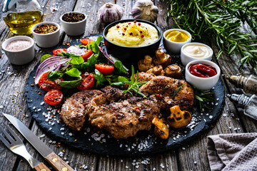 Fried pork steaks with potato puree and vegetable salad served on black stone plate on wooden table
