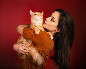 Beautiful happy smiling young woman holding on the hands and hugging with love her red maine coon kitten. Closeup portrait on red holiday background.