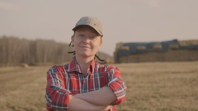 Portrait of successful woman farmer in field on background of harvested haystacks for cattle. Contented female livestock breeder smiling looks at camera.