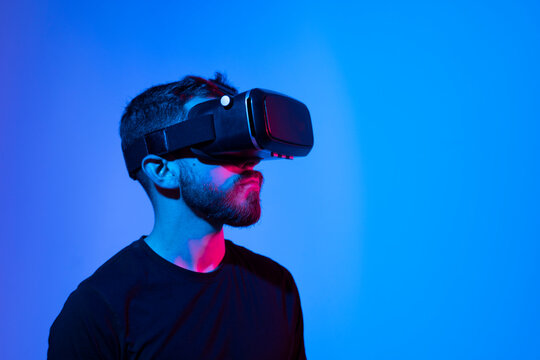 Virtual reality world. Bearded guy in vr headset, touching something while playing video game in metaverse with his friends, standing in neon light.