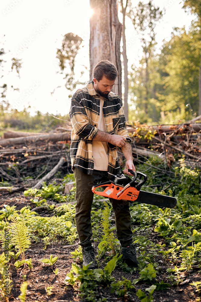 Wall mural Nice young caucasian lumberjack in casual shirt is preparing to cut trees with electric saw, standing in forest alone, posing, at summer evening. outdoor activity. strong man with saw. woodcutter.