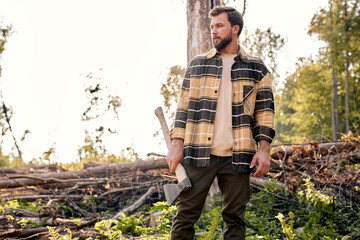 Confident lumberjack works in summer forest. Lumberjack with an ax in the evening. Wood man holding...