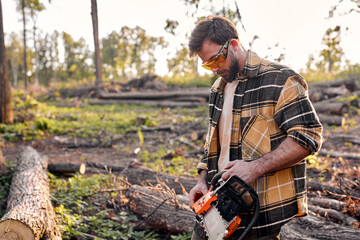 Lumberman is going to saw branches with chainsaw from felled tree in summer forest. Concept of professional logging, wood working. Young male in casual outfit preparing to chop, cut trees