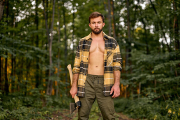 Brutal handsome european man in open plaid shirt carry large splitting axe in summer forest natural landscape, lumberman with perfect muscular body posing at camera, shirtless sexy lumberman