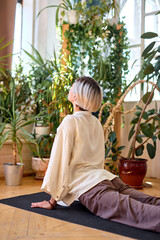 Young flexible and strong female doing fitness yoga exercises at home, on fitness mat, stretching body. Side view on slender woman in bright room full of green plants. Healthy lifestyle concept
