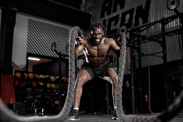 Fototapeta na wymiar afro american fit athlete doing battle ropes exercise at crossfit gym. African Male wear black shorts training with rope. sport motivation, cross fit, fitness concept. cardio training