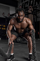 Fototapeta na wymiar Young black man doing strength training using battle ropes at modern dark gym. Athlete moving the ropes in wave motion as part of fat burning workout, active intense cross fit training, exercising