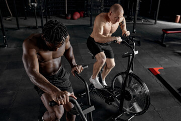 Fototapeta na wymiar Men exercise bike gym cycling training fitness. Two Fitness male using air bike cardio workout. Two Athlete guys with naked torso biking indoor at gym, exercising legs. Cross functional training.