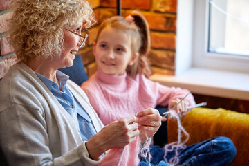 Grandmother is knitting sitting on sofa, cute little daughter is learning to knit sitting next to senior granny. Side view on friendly aged lady with daughter. Hobby, leisure, weekends