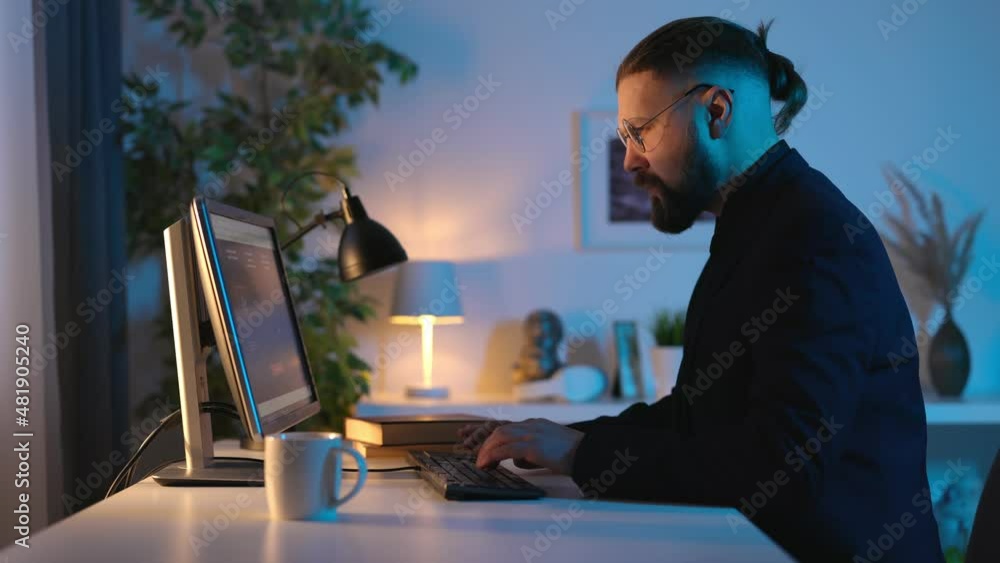 Poster side view of bearded caucasian man sitting at desk during evening time and typing on modern computer