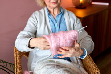 cropped older caucasian female is knitting alone at home, creating clothes, holding half-maked scarf, sitting in cozy room in domestic atmosphere, spending weekend, leisure time, relax, hobby