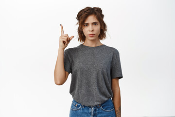 Confused young woman asks question when points finger up, see smth strange, looks concerned when...