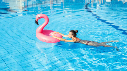 Summer pool relaxing. Young sexy woman in bikini swimsuit float with pink flamingo in blue water....