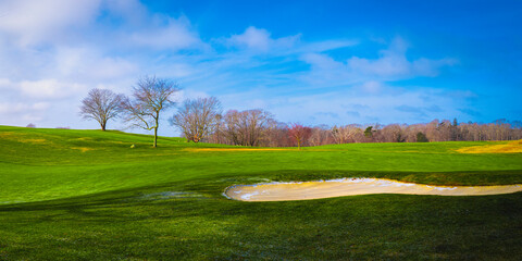 Tranquil rolling green hills with bare trees of the forest and soft floating white clouds in the blue sky.