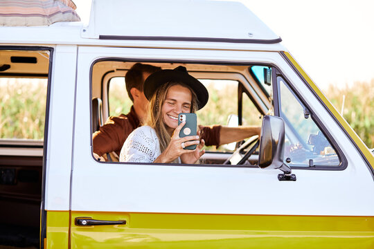 Young caucasian couple on road during travel in countryside, take photo of nature landscape using smartphone, enjoy adventure together, laughing, man driving yellow retro style mini van. side view