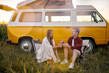 Beautiful happy Hippie couple sitting next to yellow retro style van vehicle camper trailer, caucasian man and woman drinking cup of tea from thermos. Female camping, traveling, hitchhiking.