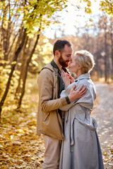 Fototapeta premium young and attractive couple hugging with love in the middle of park during autumn at sunny day, caucasian man and woman in coats feel tenderness to each other, good relationships concept