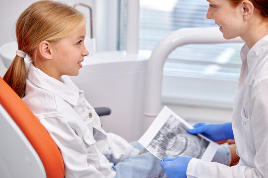 Side View On Kid Girl Listening To Female Dentist, Having Talk About Teeth Treatment. Stomatologist Showing X-ray Picture To Child Girl Patient. Medicine, healthcare, treatment concept