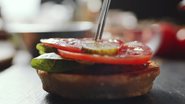 Close up shot of putting slice of dill pickle with tweezers on burger bun with lettuce and tomato